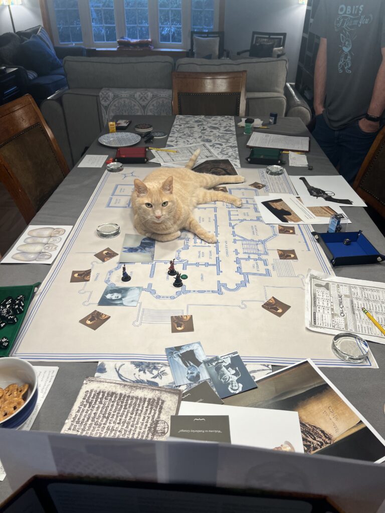 An orange cat sitting int the middle of a table on top of maps and materials used in a Call of Cthulhu tabletop role playing game
